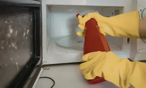 Manos con guantes amarillos rociando el interior de un microondas sucio con un limpiador.