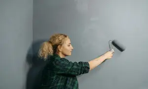 Mujer rubia con coleta pinta pared gris con rodillo. Se pregunta cuanto cuesta pintar un piso.