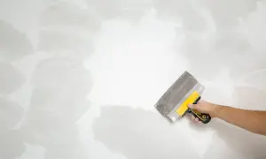 Mano con espátula amarilla y gris trabajando en una pared, alisando la textura para como quitar gotele.