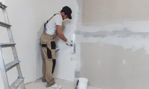 Obrero aplica yeso para pared con llana. Escalera y cubo de material a un lado.
