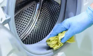 Mano con guante azul limpiando el borde de la lavadora con un paño amarillo. Un chorrito de vinagre de limpieza lavadora puede ayudar a desinfectar.