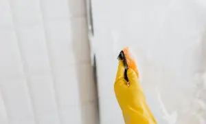 Mano con guante amarillo limpiando mampara de ducha con esponja.