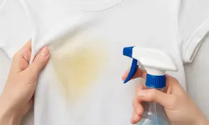 Manos rociando un quitamanchas en una camiseta blanca con una mancha amarilla. Aprende como quitar manchas amarillas en seco.