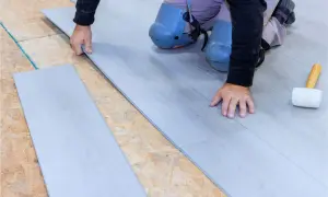 Man con rodilleras coloca cuidadosamente tablas de suelo laminado gris sobre una base de madera.