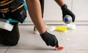 Persona con guantes negros frota las juntas de los azulejos con un cepillo naranja, aplicando un producto para blanquear juntas azulejos.