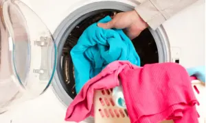 Mano mete ropa azul en lavadora. La ropa rosa y azul en la cesta sugieren que la lavadora huele mal.