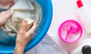 Manos lavando ropa en un barreño azul con jabón, para saber como quitar resina de la ropa. Cerca, un recipiente con detergente en polvo.