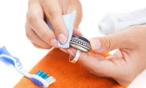 Manos limpiando un anillo de plata con un paño azul. Se ve un cepillo de dientes y una pasta de dientes, indicando cómo se limpia la plata en casa.