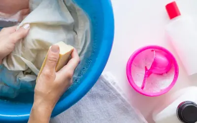 Manos lavando ropa en un barreño azul con jabón, para saber como quitar resina de la ropa. Cerca, un recipiente con detergente en polvo.