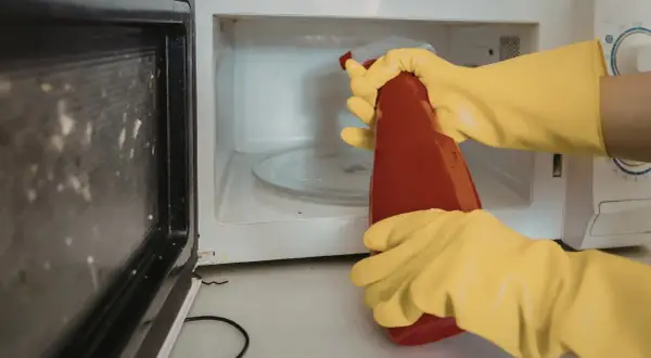 Manos con guantes amarillos rociando el interior de un microondas sucio con un limpiador.
