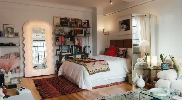 Un dormitorio con un diseño estudio moderno. Cama blanca, estanterías llenas de libros, un espejo ondulado y alfombras coloridas.