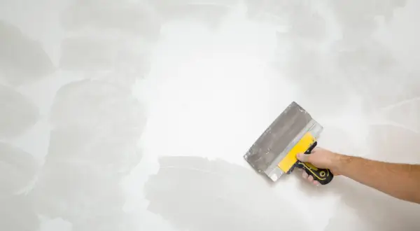 Mano con espátula amarilla y gris trabajando en una pared, alisando la textura para como quitar gotele.