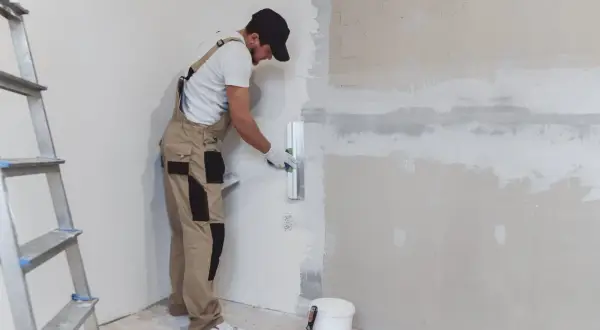 Obrero aplica yeso para pared con llana. Escalera y cubo de material a un lado.