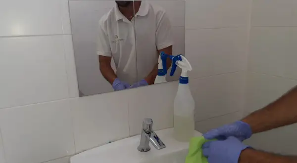 Man con guantes limpia lavabo con paño verde. Spray de limpieza cerca. Aprende como limpiar azulejos baño.