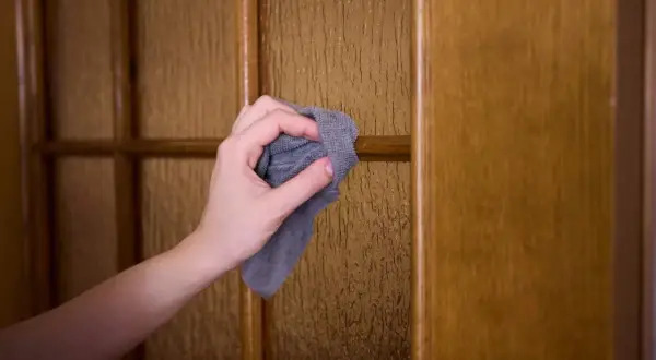 Mano limpiando con paño gris una puerta de madera con cristal texturizado. Aprende como limpiar las puertas de madera fácilmente.