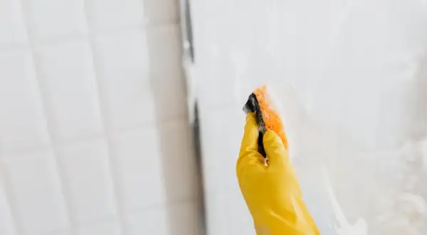 Mano con guante amarillo limpiando mampara de ducha con esponja.