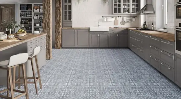 Cocina moderna con suelo hidráulico de diseño geométrico en tonos grises y blancos. Muebles de cocina grises, encimera de madera y barra americana con taburetes.