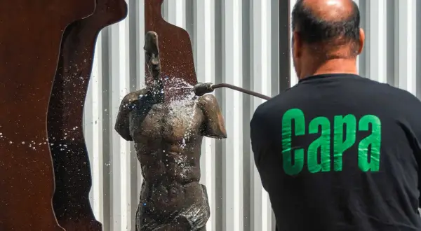 Hombre con camiseta negra con letras verdes "Capa" limpia una estatua de bronce con agua a presión.