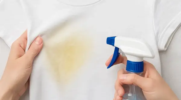 Manos rociando un quitamanchas en una camiseta blanca con una mancha amarilla. Aprende como quitar manchas amarillas en seco.