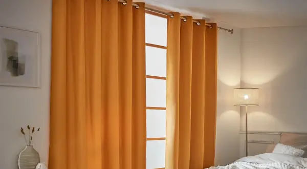 Cortinas aislantes naranjas cubren una ventana, creando un ambiente acogedor. A la izquierda, un cuadro abstracto y un jarrón con espigas.