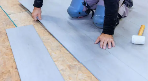 Man con rodilleras coloca cuidadosamente tablas de suelo laminado gris sobre una base de madera.