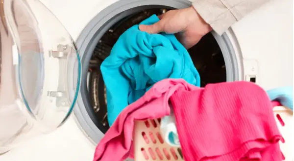 Mano mete ropa azul en lavadora. La ropa rosa y azul en la cesta sugieren que la lavadora huele mal.