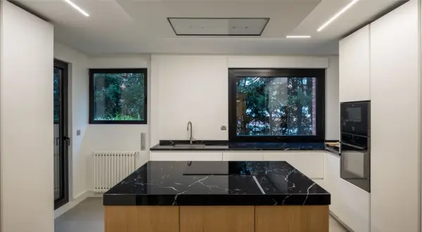Moderna cocina con isla central de mármol negro y madera. Encimeras a juego, electrodomésticos integrados y grandes ventanales.