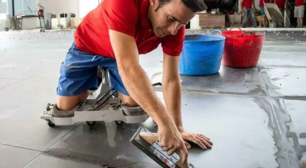 Un trabajador con rodilleras aplica cemento para saber como poner baldosas en el suelo.