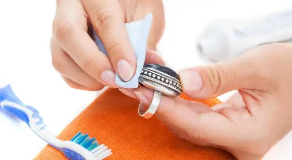Manos limpiando un anillo de plata con un paño azul. Se ve un cepillo de dientes y una pasta de dientes, indicando cómo se limpia la plata en casa.