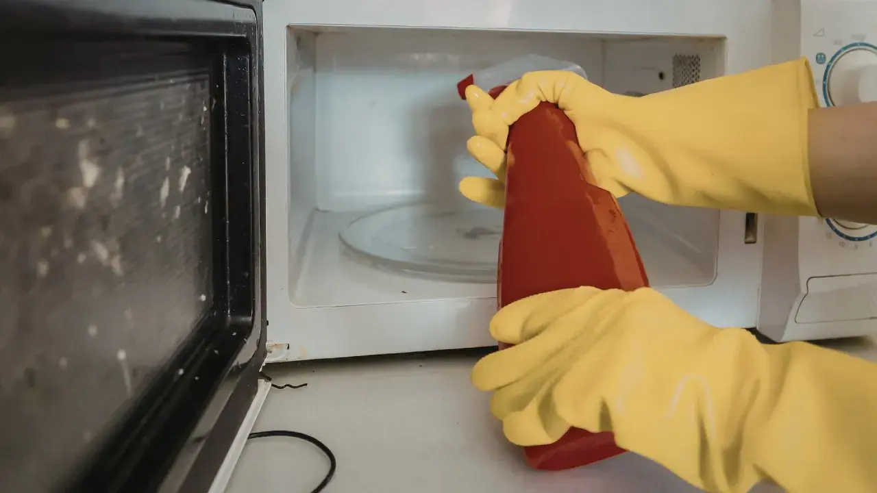 Manos con guantes amarillos rociando el interior de un microondas sucio con un limpiador.