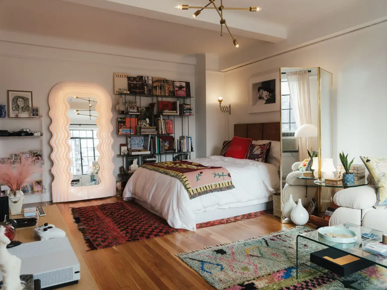 Un dormitorio con un diseño estudio moderno. Cama blanca, estanterías llenas de libros, un espejo ondulado y alfombras coloridas.