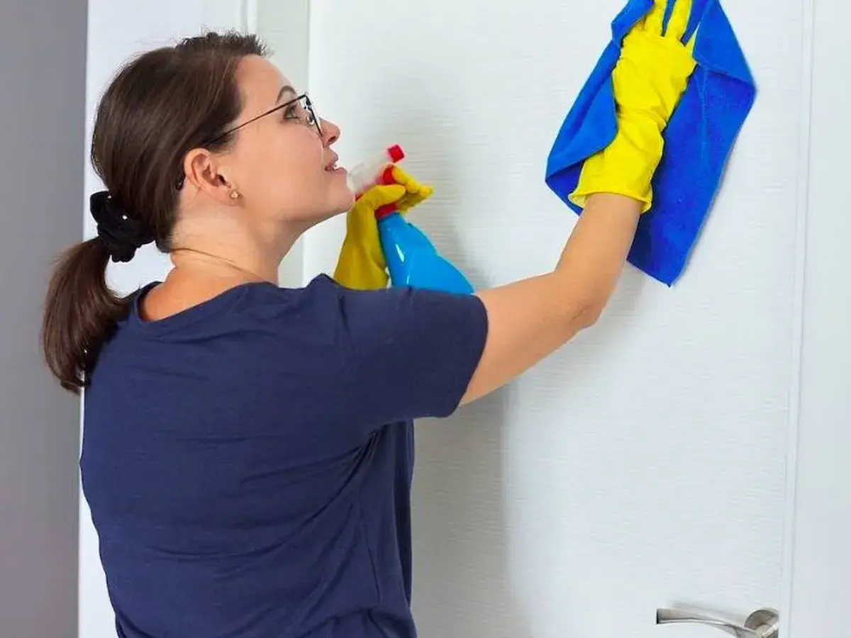Mujer con guantes amarillos y paño azul limpiando puerta lacada en blanco con spray.