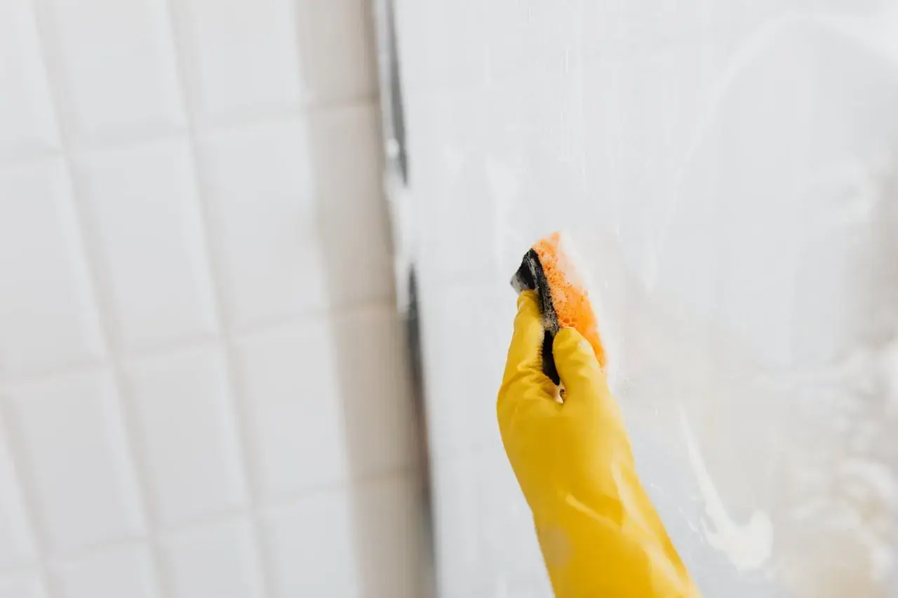 Mano con guante amarillo limpiando mampara de ducha con esponja.