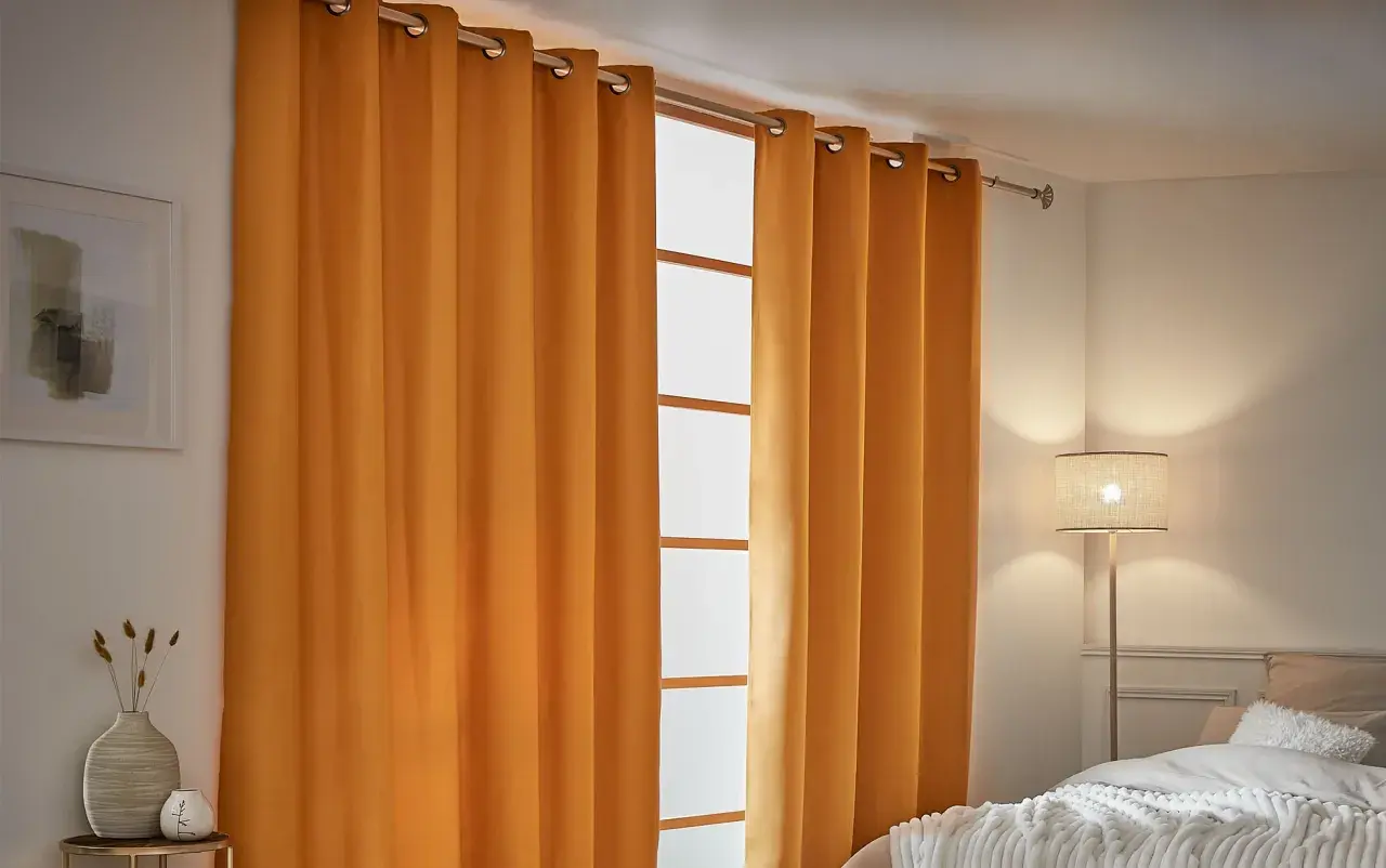 Cortinas aislantes naranjas cubren una ventana, creando un ambiente acogedor. A la izquierda, un cuadro abstracto y un jarrón con espigas.