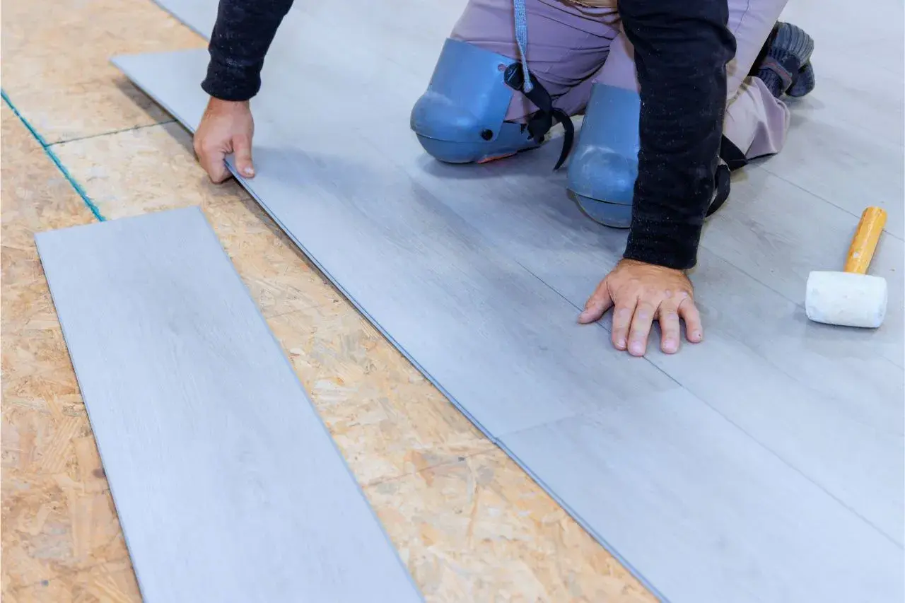 Man con rodilleras coloca cuidadosamente tablas de suelo laminado gris sobre una base de madera.