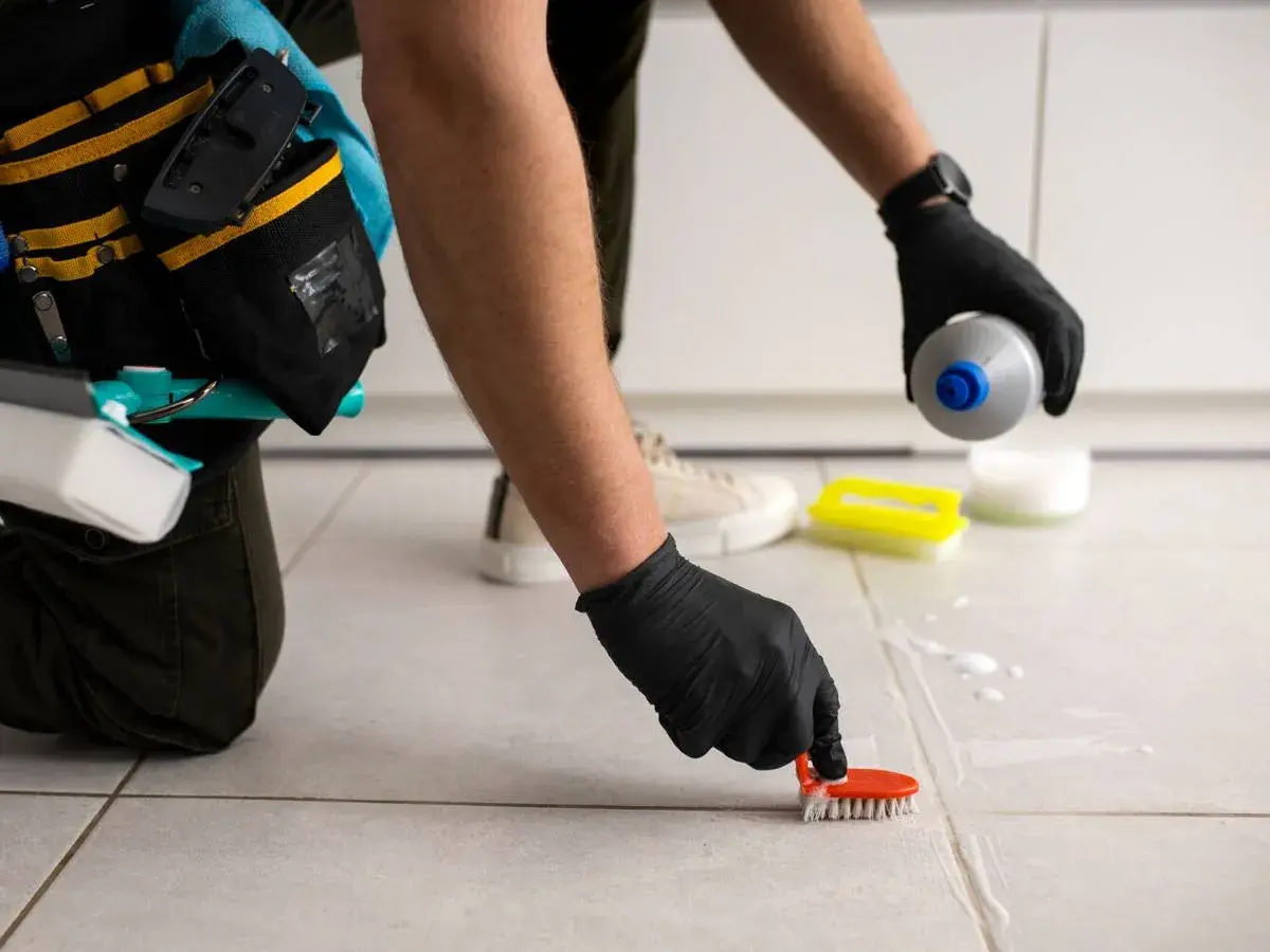 Persona con guantes negros frota las juntas de los azulejos con un cepillo naranja, aplicando un producto para blanquear juntas azulejos.
