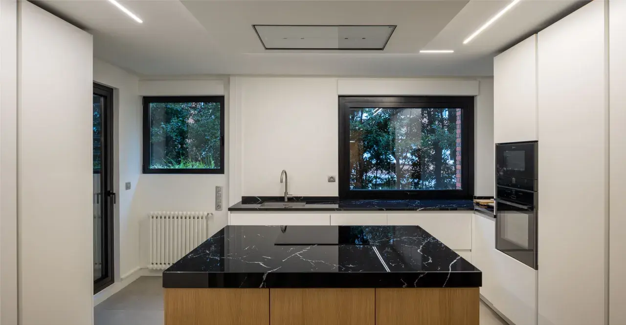 Moderna cocina con isla central de mármol negro y madera. Encimeras a juego, electrodomésticos integrados y grandes ventanales.