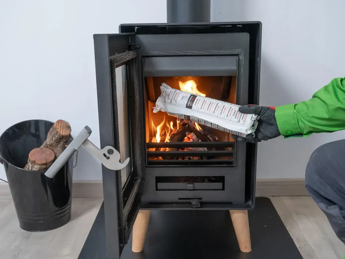 Persona con guantes verdes y grises introduce un producto envasado en una chimenea encendida. Es parte de la limpieza de chimeneas.