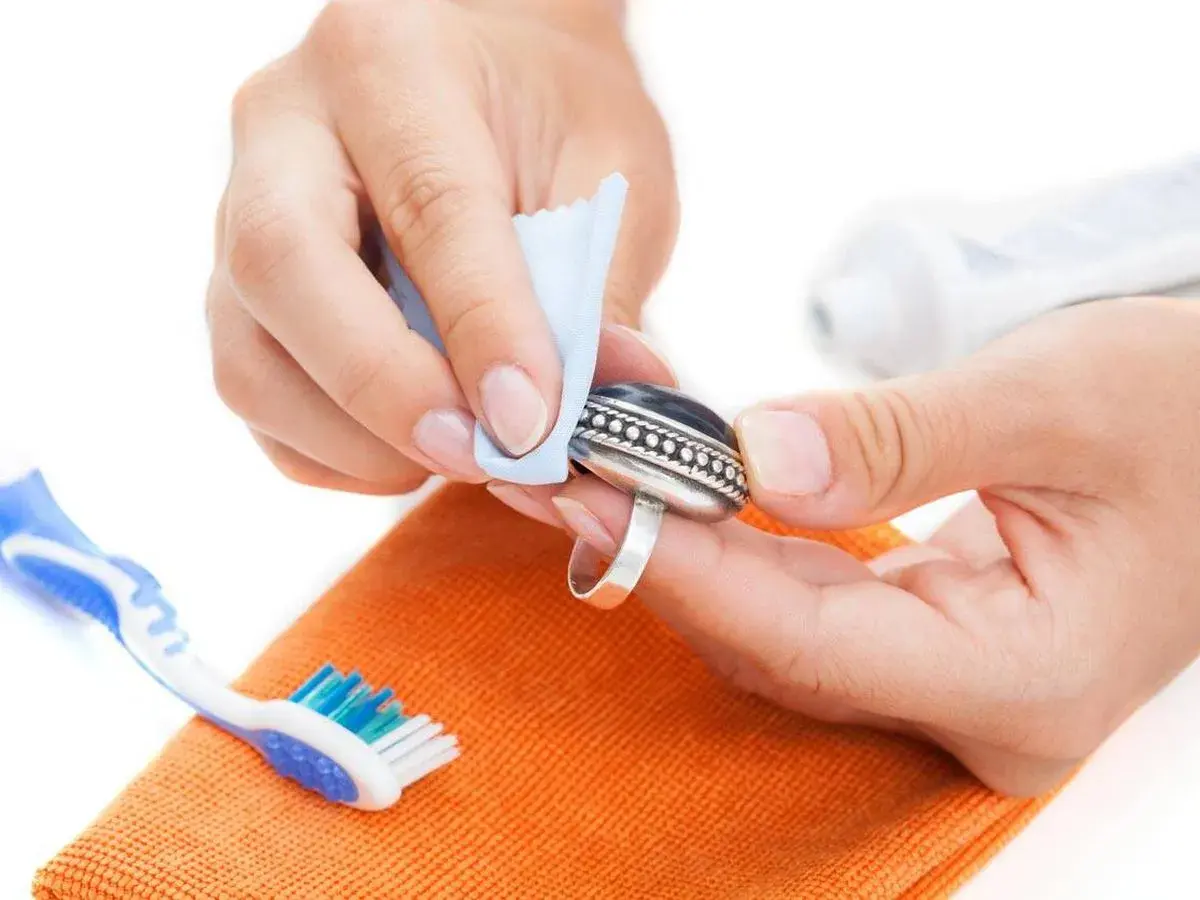 Manos limpiando un anillo de plata con un paño azul. Se ve un cepillo de dientes y una pasta de dientes, indicando cómo se limpia la plata en casa.