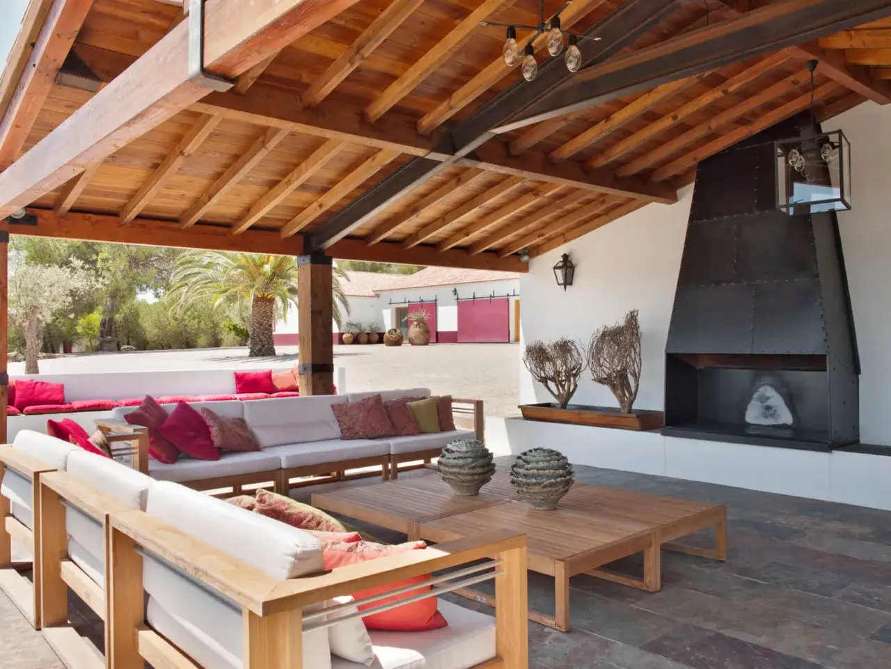 Amplia terraza con sofás blancos, mesas de madera y chimenea metálica, bajo techos de madera rústicos.