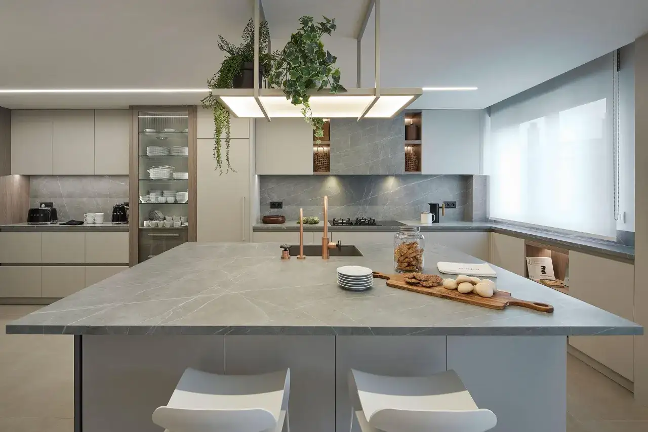 Moderna cocina con isla central de mármol gris. Encimeras a juego, armarios minimalistas y grifería de cobre. Plantas colgantes añaden un toque verde.