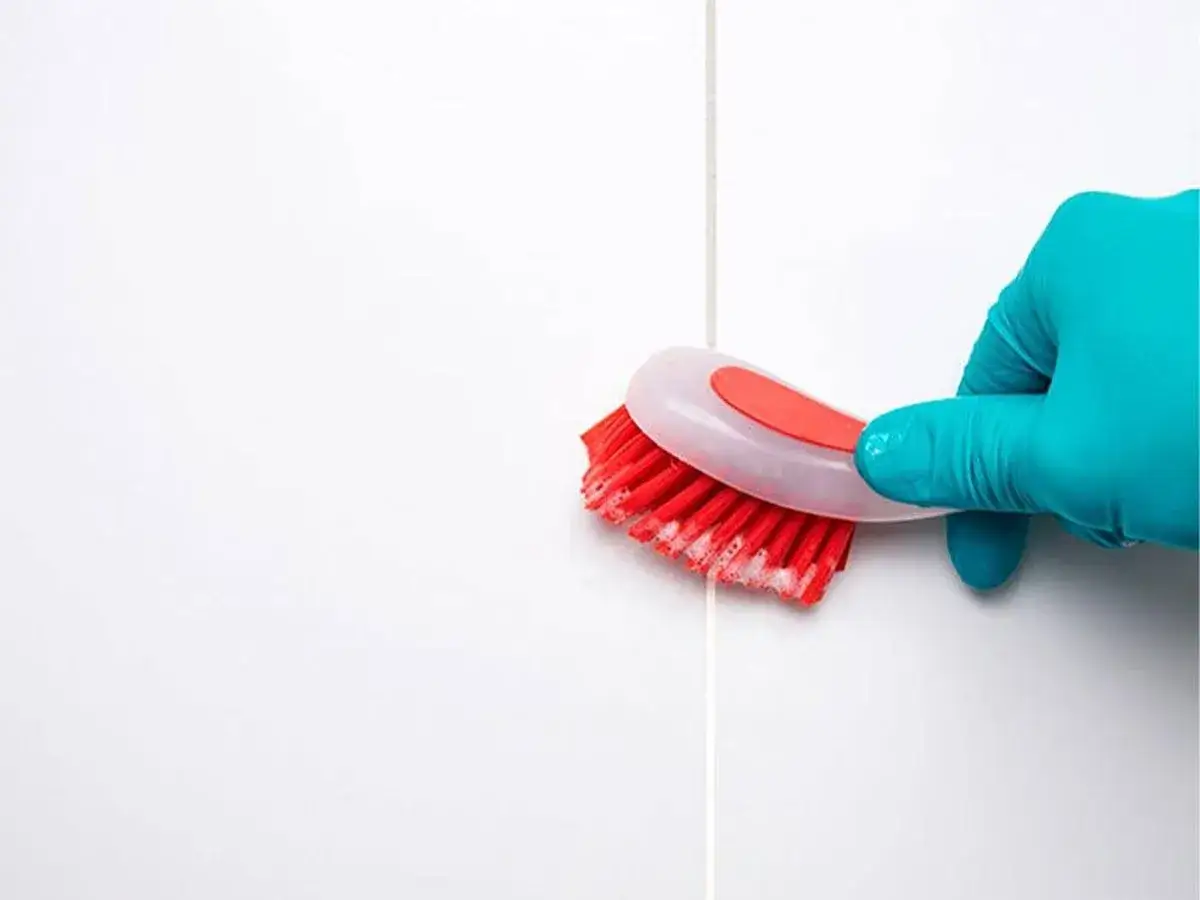 Mano con guante azul cepillando juntas de azulejos blancos con un cepillo rojo y espuma, para blanquear juntas azulejos.