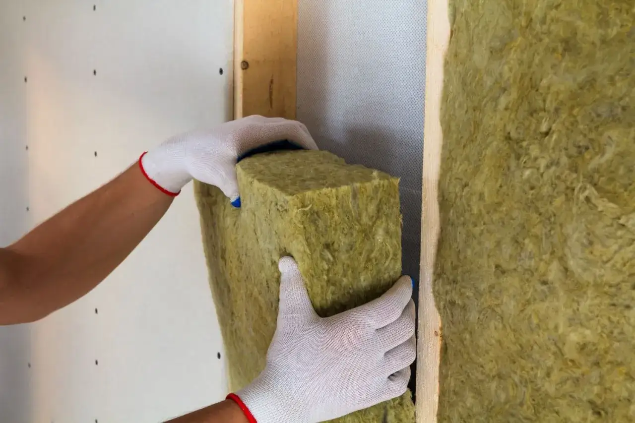 Manos enguantadas colocan material aislante en una pared, mejorando el aislamiento del suelo.