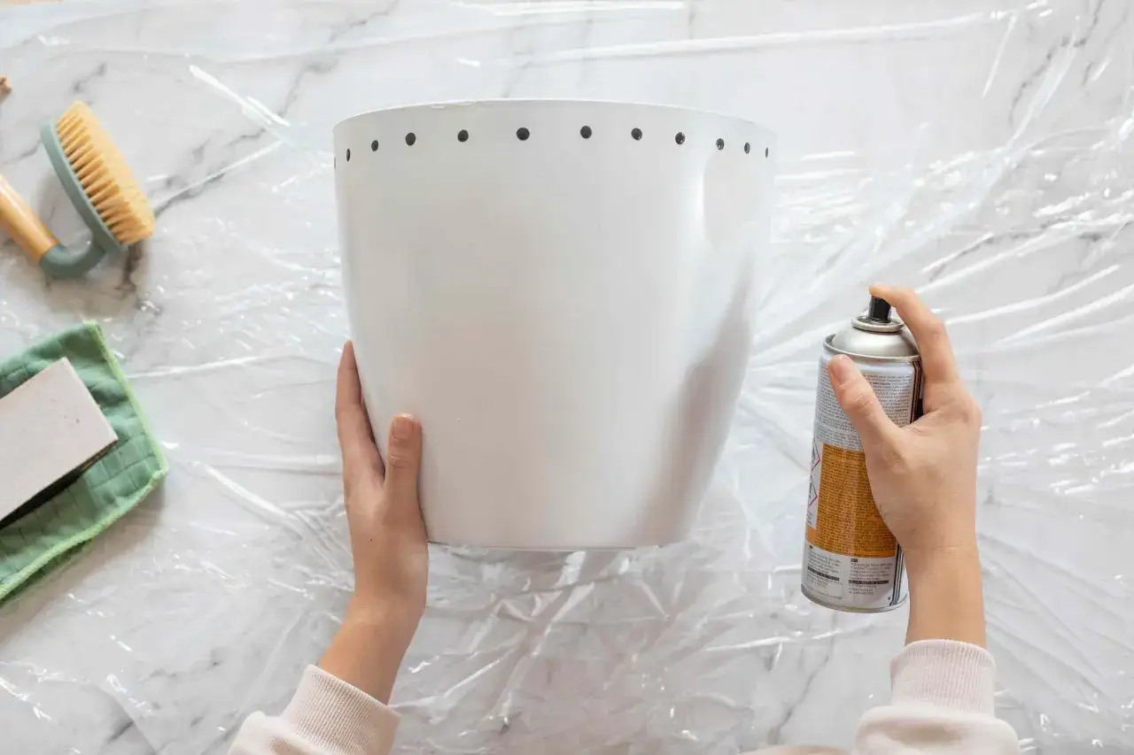 Manos rociando pintura en spray sobre una maceta blanca, preparándola para un proyecto de como pintar plastico.