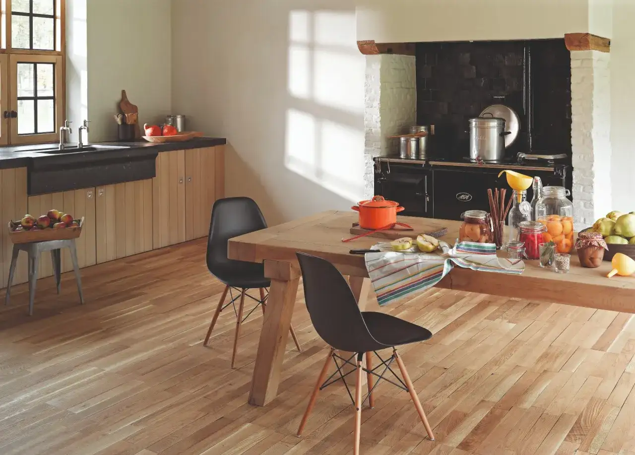 Cocina rústica con suelo de madera natural, mostrando diferentes tipos de parquet. Mesa de madera con sillas negras, cocina de leña y encimera con conservas.