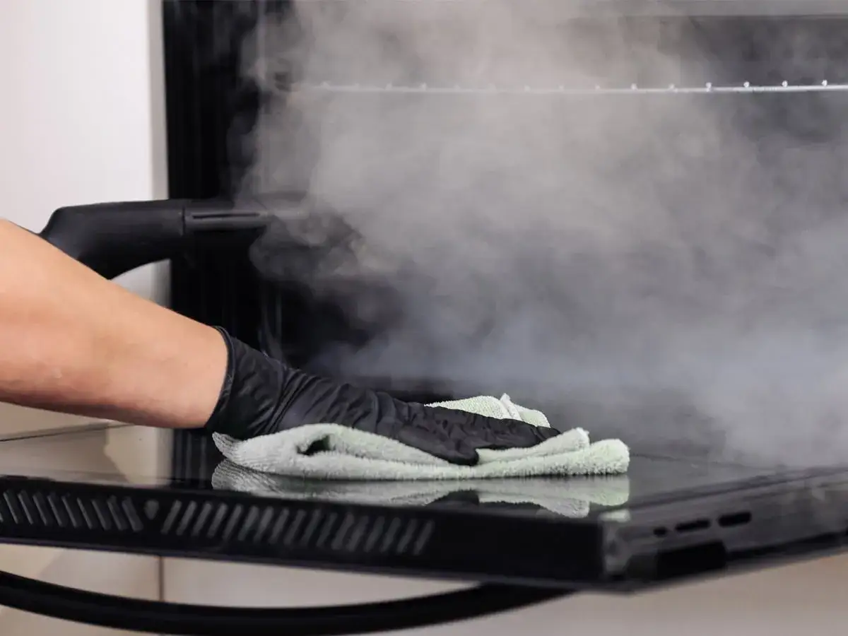 Mano enguantada usa vapor para limpiar horno. El vapor disuelve la grasa, facilitando la limpieza.