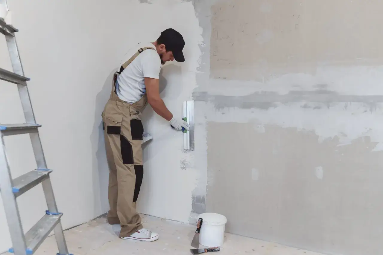 Obrero aplica yeso para pared con llana. Escalera y cubo de material a un lado.