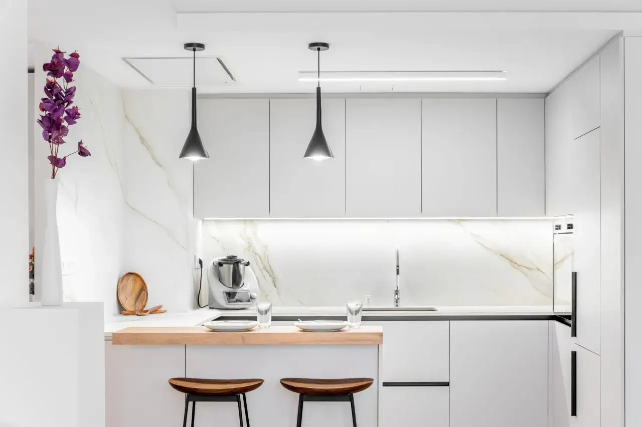 Cocina moderna y luminosa, ideal para reformas de cocinas pequeñas. Destacan los muebles blancos, la encimera de mármol y la barra de madera con taburetes.