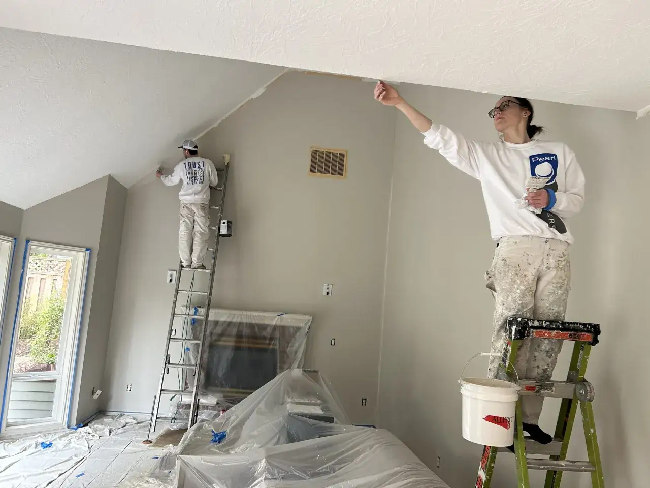 Dos pintores trabajan en una habitación, uno en una escalera alta y otro en una más baja. Ambos pintan el techo, respondiendo a la pregunta de si se pinta primero el techo o las paredes.