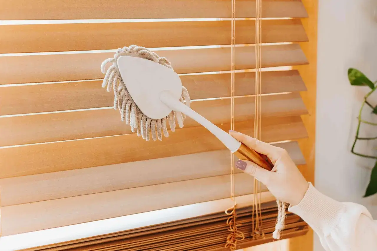 Mano limpiando persianas de madera con un plumero. Un truco sencillo para saber como limpiar persianas y mantener tu hogar impecable.